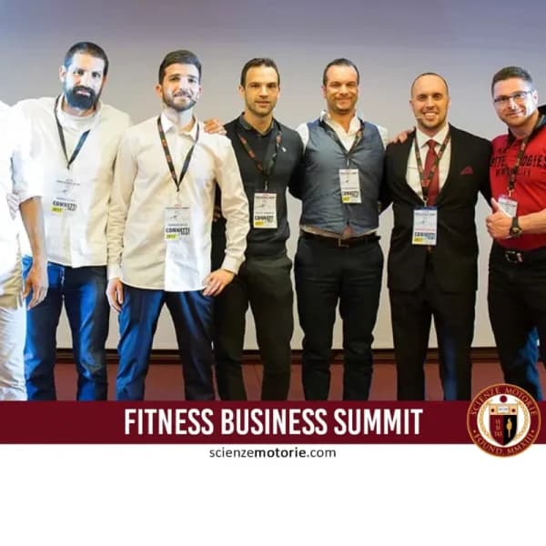 Otto persone in piedi su un palco durante un evento, indossano abiti formali con badge al collo.