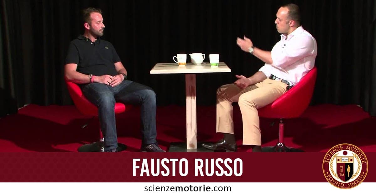 Due uomini seduti su poltrone rosse davanti a un tavolo con tazze e bicchiere, uno con camicia bianca e pantaloni chiari, l’altro con maglietta nera e jeans, in una stanza con pavimento e sfondo scuri. Scritta "Fausto Russo" e logo Scienze Motorie in basso.