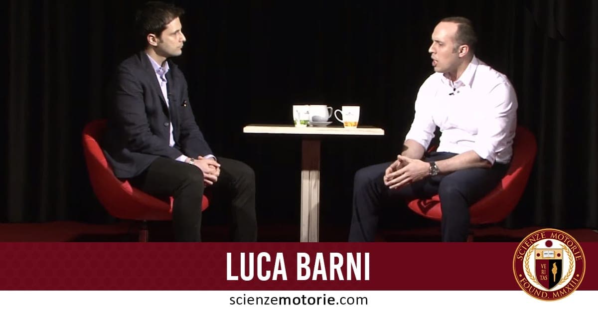 Due uomini vestiti formalmente seduti su sedie rosse di fronte a un tavolo con tazze, con sfondo scuro; Luca Barni.