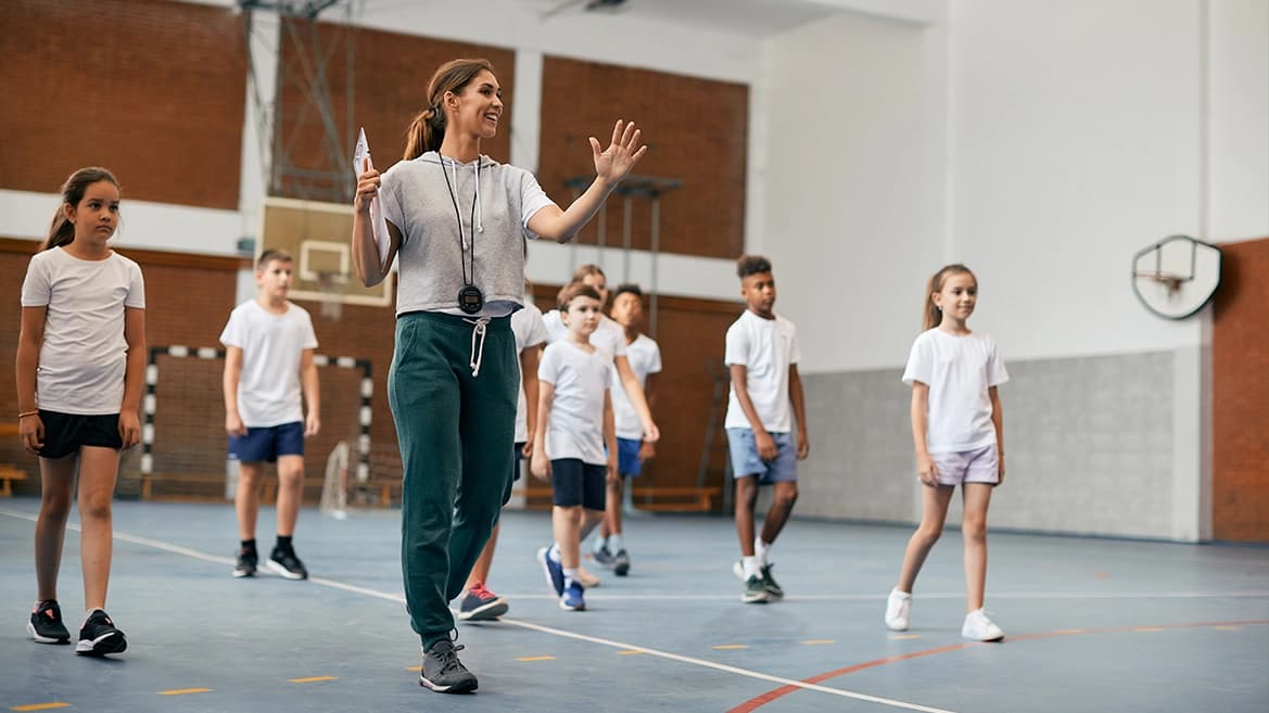 Insegnante di educazione fisica con cronometro che guida un gruppo di bambini in palestra scolastica con canestro da basket, logo scienzemotorie.com visibile.